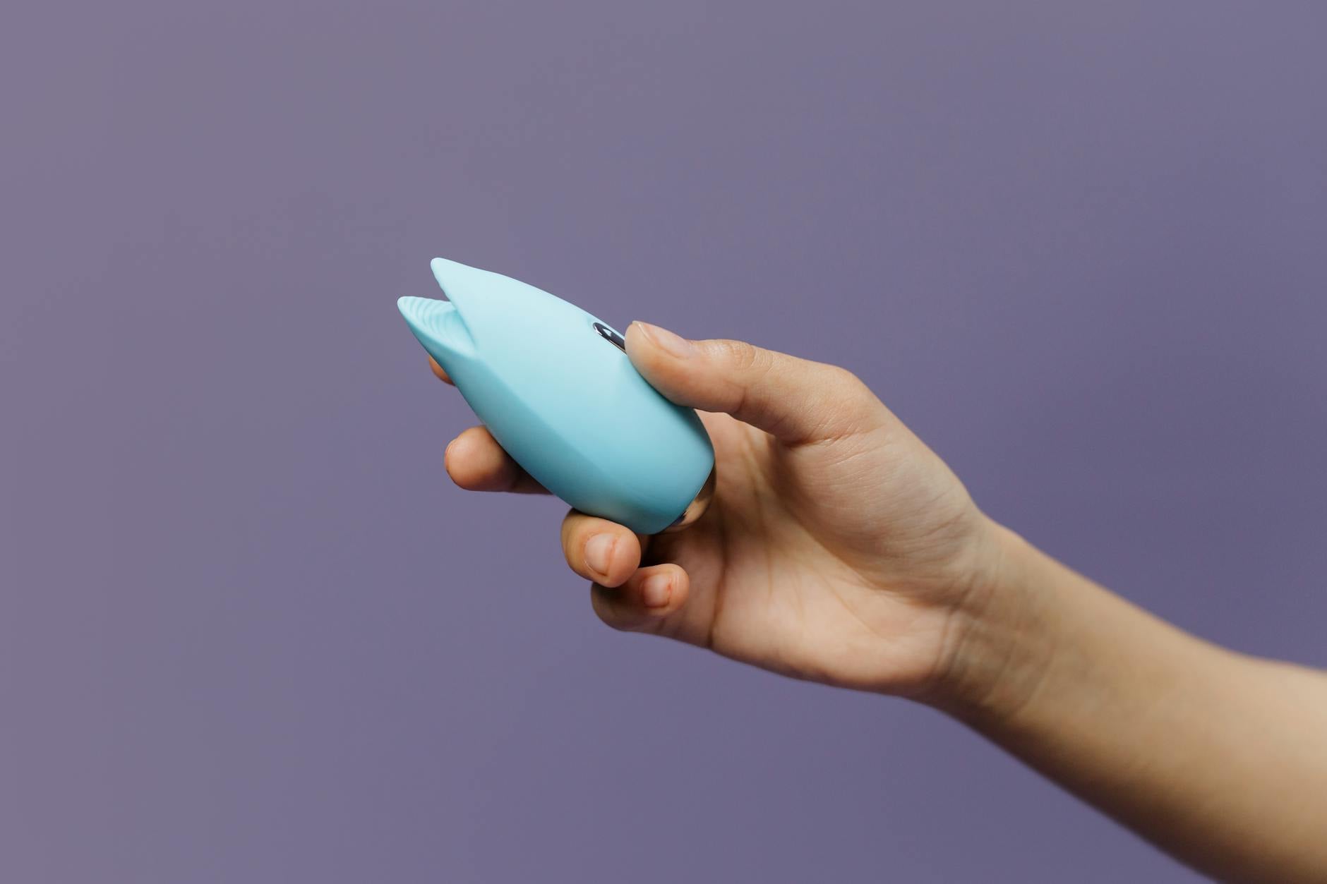 A blue silicone clitoral vibrator held in hand against a purple background, representing external pleasure without penetration