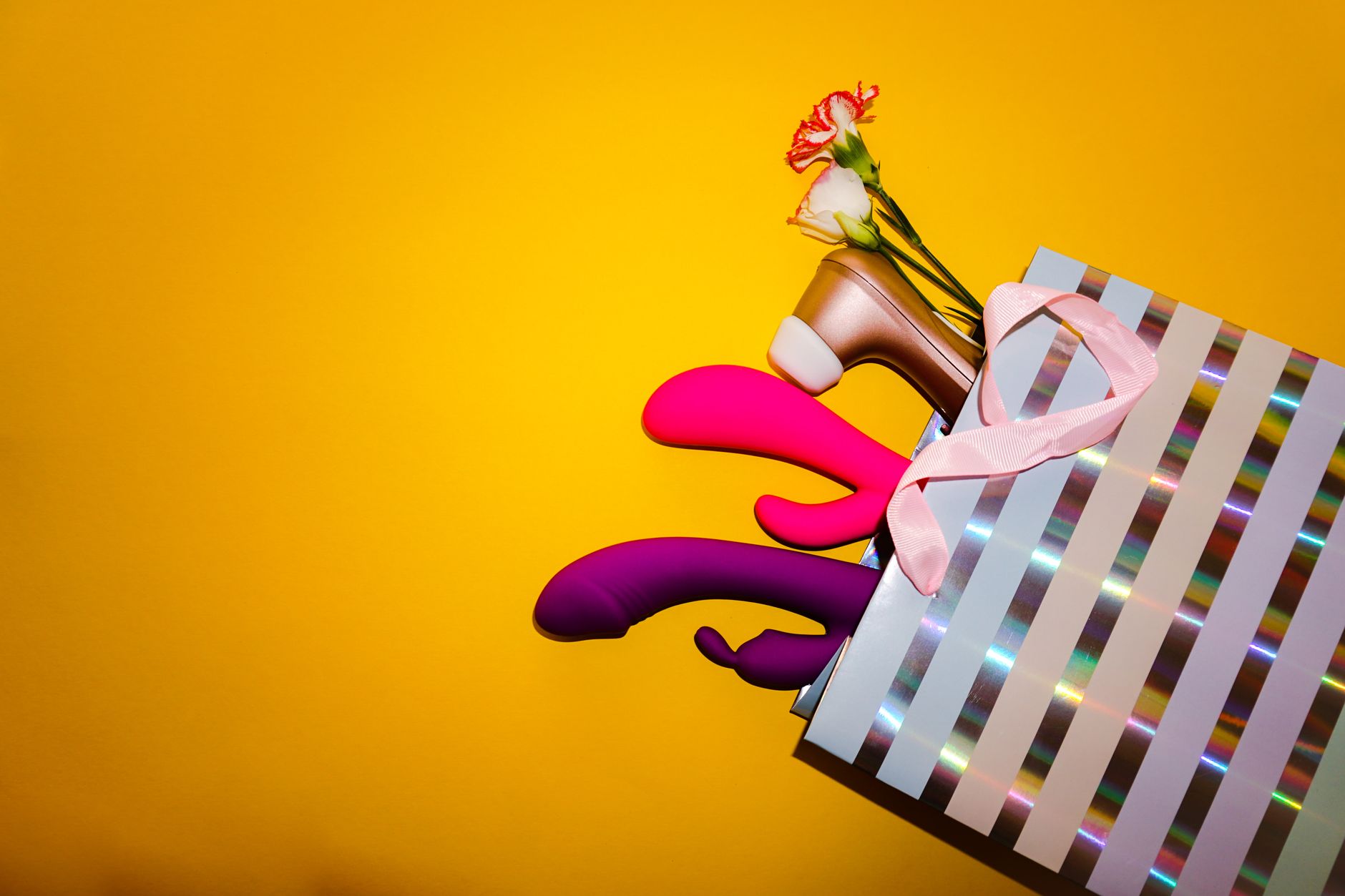 Colorful adult vibrators and toys displayed with flowers in a holographic gift bag on bold yellow background
